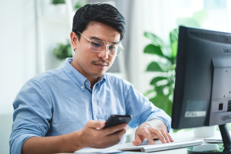 A man wearing glasses is sitting at a desk with a computer monitor and a cell phone. He is looking at the cell phone while typing on the computer